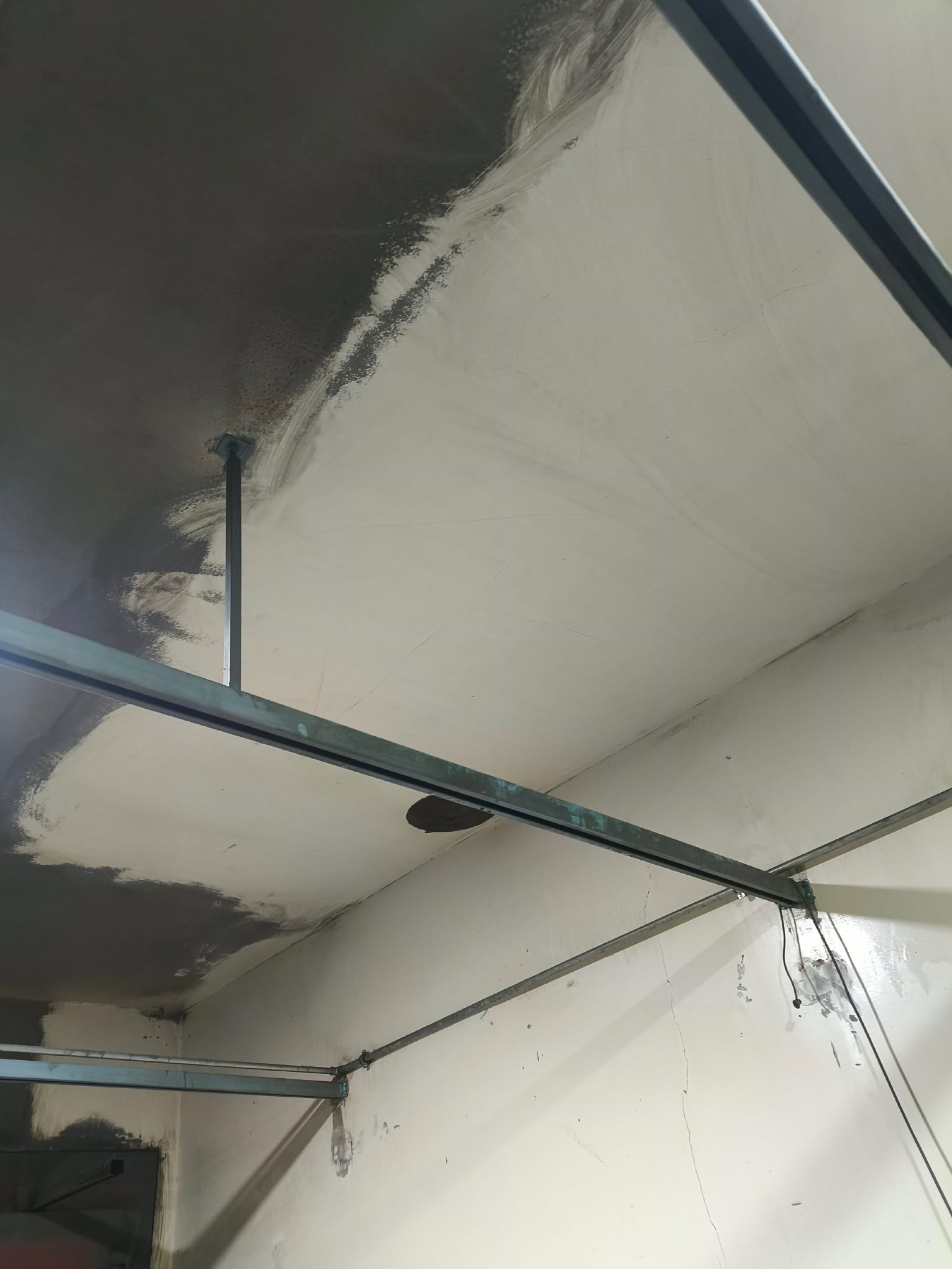 Malowanie sufitu w trakcie prac. Widoczne metalowe profile podwieszane i fragmenty sufitu pokryte szarą farbą, kontrastujące z jasną, niemalowaną powierzchnią.