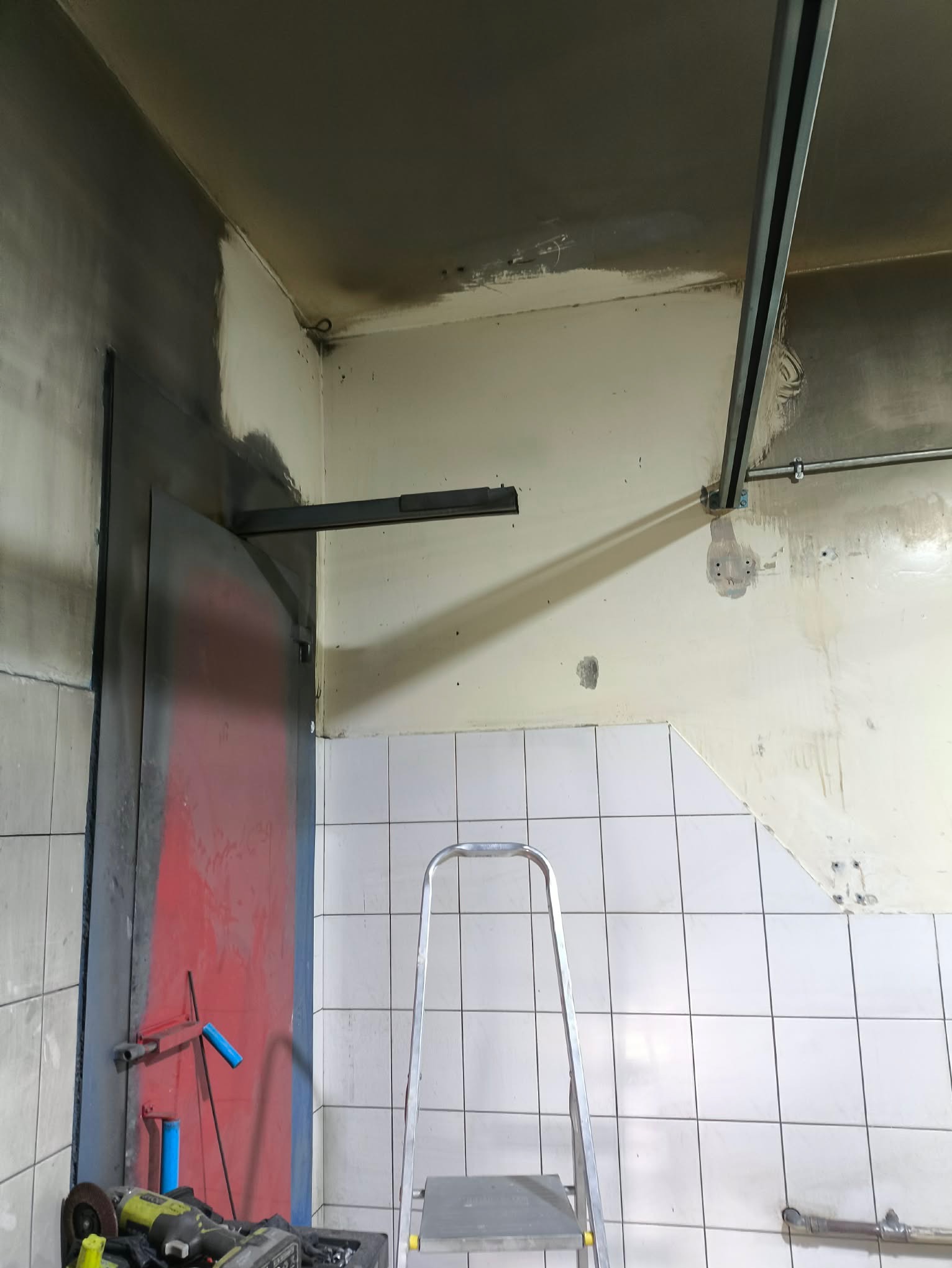 Zniszczone pomieszczenie po pożarze z widocznymi śladami okopcenia na ścianach i suficie, drabina i narzędzia na podłodze, ukazujące etap sprzątania po pożarze.