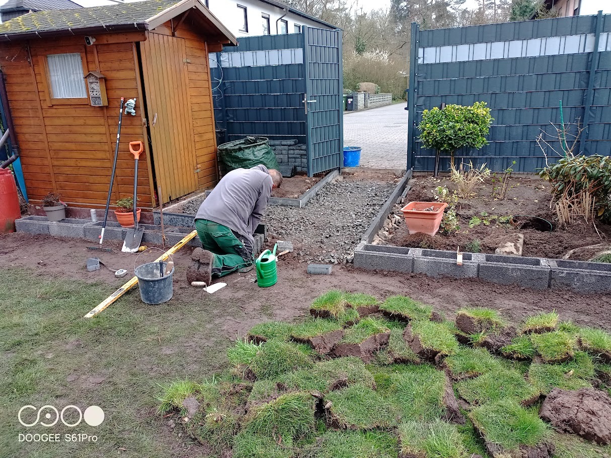 Układanie obrzeży z betonowych bloczków w ogrodzie. Widoczne narzędzia, trawa w rolkach i przygotowane podłoże. Prace ogrodowe w toku, zielony trawnik w budowie.