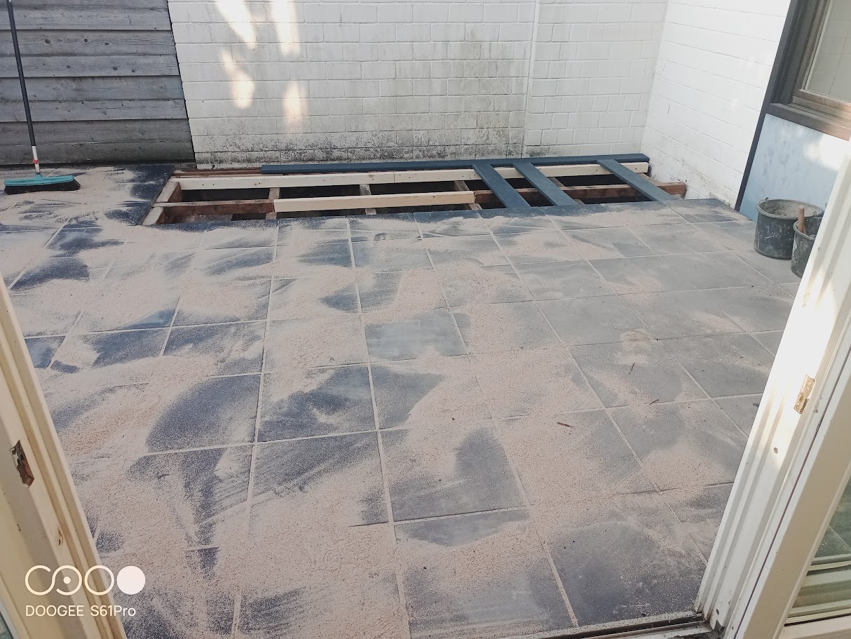 Przygotowanie podłoża pod taras: ułożone legary na betonowej posadzce, zasypane piaskiem. Widoczna ściana z cegły i fragment drzwi balkonowych.