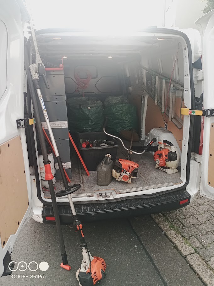 Wnętrze białego busa z otwartymi drzwiami, wypełnione narzędziami ogrodniczymi: kosiarki, nożyce do żywopłotu, worki na odpady, drabina. Widok z tyłu pojazdu.