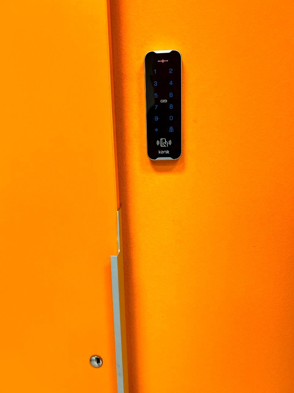 Pomarańczowy panel dotykowy z klawiaturą numeryczną i czytnikiem kart zbliżeniowych, zamontowany na pomarańczowej ścianie obok drzwi z metalowym wykończeniem.