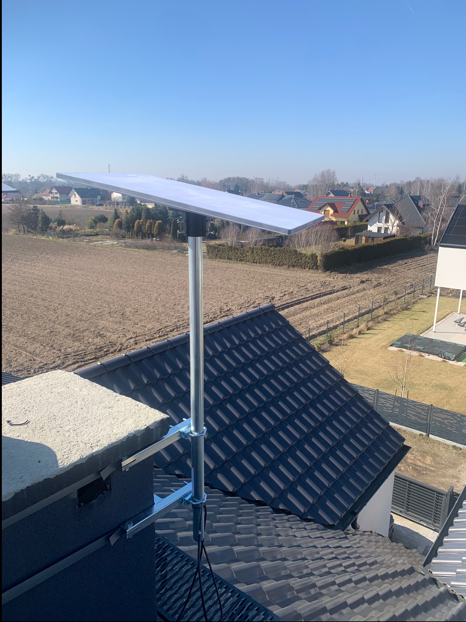 Antena satelitarna zamontowana na kominie domu jednorodzinnego z widokiem na okolicę w słoneczny dzień. Widoczne kable antenowe.