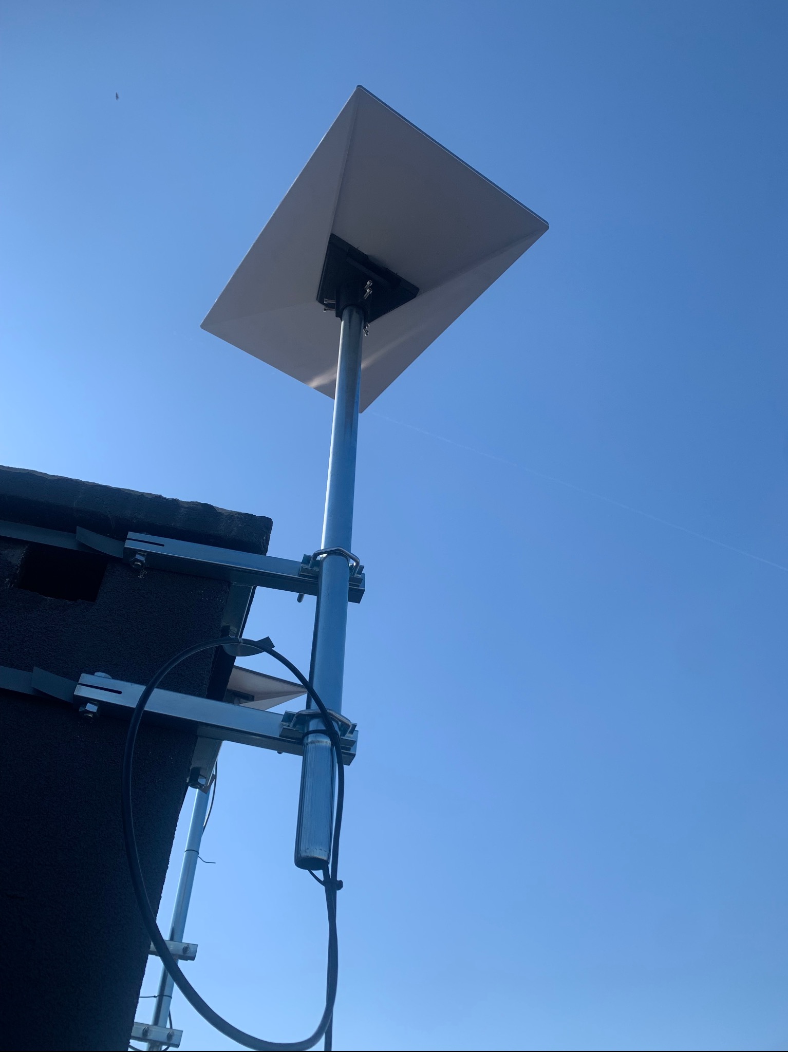 Antena satelitarna zamontowana na dachu budynku, widoczna na tle błękitnego nieba. Metalowa konstrukcja wspornika, kabel antenowy. Ujęcie z dołu.