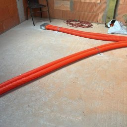 KWD-INSTAL SPÓŁKA Z OGRANICZONĄ ODPOWIEDZIALNOŚCIĄ - Pomarańczowe rury karbowane ułożone na surowym betonie w pomieszczeniu z ceglanymi ścianami. Widoczny fragment instalacji i krzesło w tle.