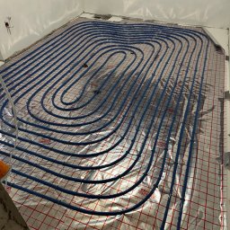 KWD-INSTAL SPÓŁKA Z OGRANICZONĄ ODPOWIEDZIALNOŚCIĄ - Niebieskie rury ogrzewania podłogowego ułożone spiralnie na srebrnej folii z czerwoną siatką w jasnym pomieszczeniu. Widoczne fragmenty ścian i podłoża w trakcie instalacji.