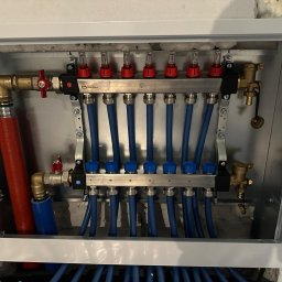 KWD-INSTAL SPÓŁKA Z OGRANICZONĄ ODPOWIEDZIALNOŚCIĄ - Rozdzielacz ogrzewania podłogowego w metalowej szafce, z widocznymi niebieskimi rurami i elementami regulacyjnymi. Widok z góry, instalacja w trakcie montażu.