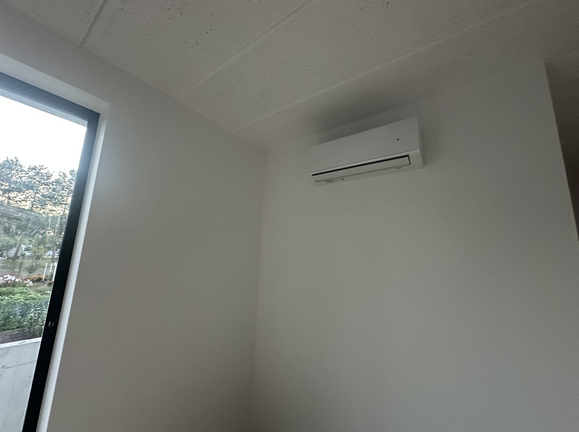 Biała klimatyzacja ścienna w jasnym wnętrzu, widok z dołu, z oknem po lewej stronie kadru. Minimalistyczny design, świeżo pomalowane ściany.