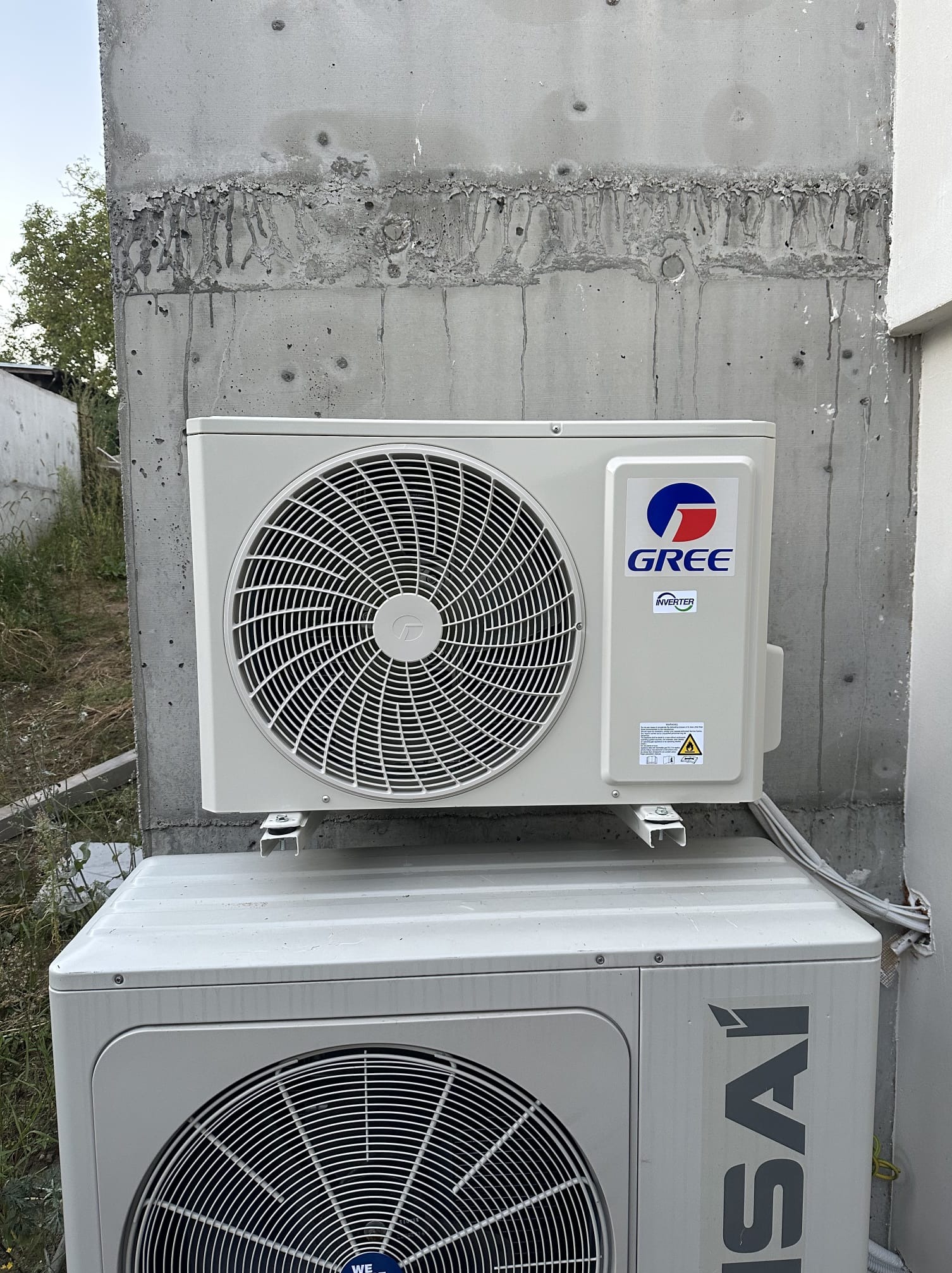 Dwa klimatyzatory zewnętrzne, marki Gree i Kaisai, zamontowane na szarej, betonowej ścianie budynku. Widoczne przewody i elementy instalacji.