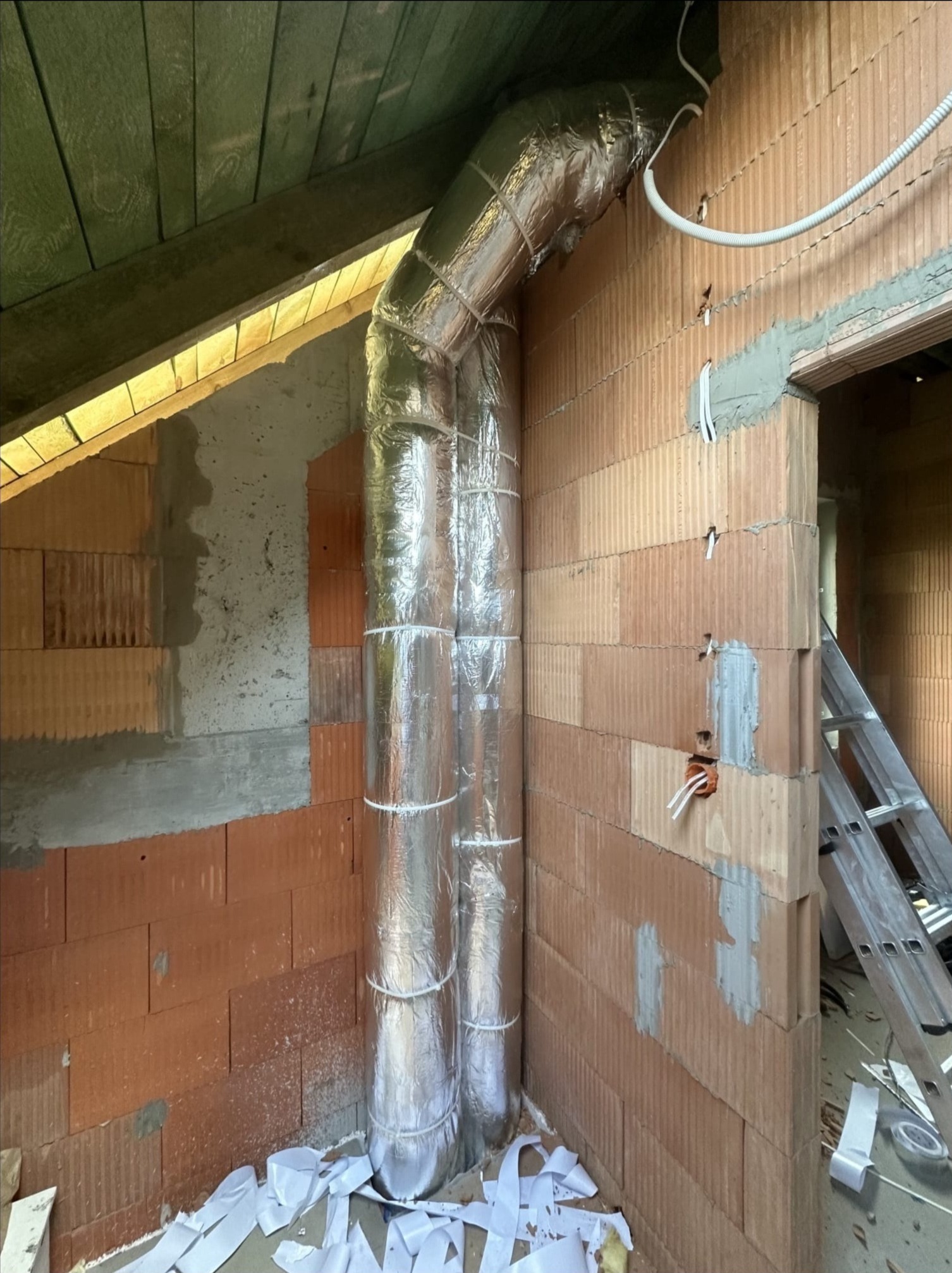 Izolowany kanał wentylacyjny z folii aluminiowej w surowym wnętrzu z cegły, widoczne przewody elektryczne i drabina. Etap instalacji rekuperacji w domu.