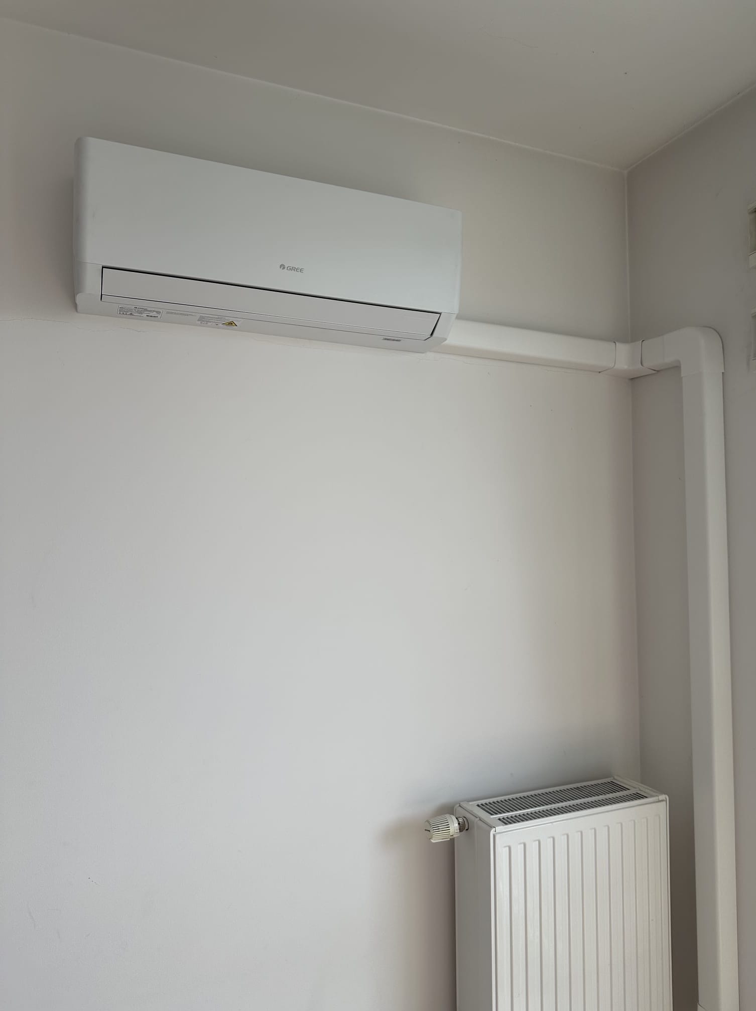 Biały klimatyzator marki Gree zamontowany na ścianie, z widocznym grzejnikiem pod spodem. Instalacja poprowadzona w białych listwach.
