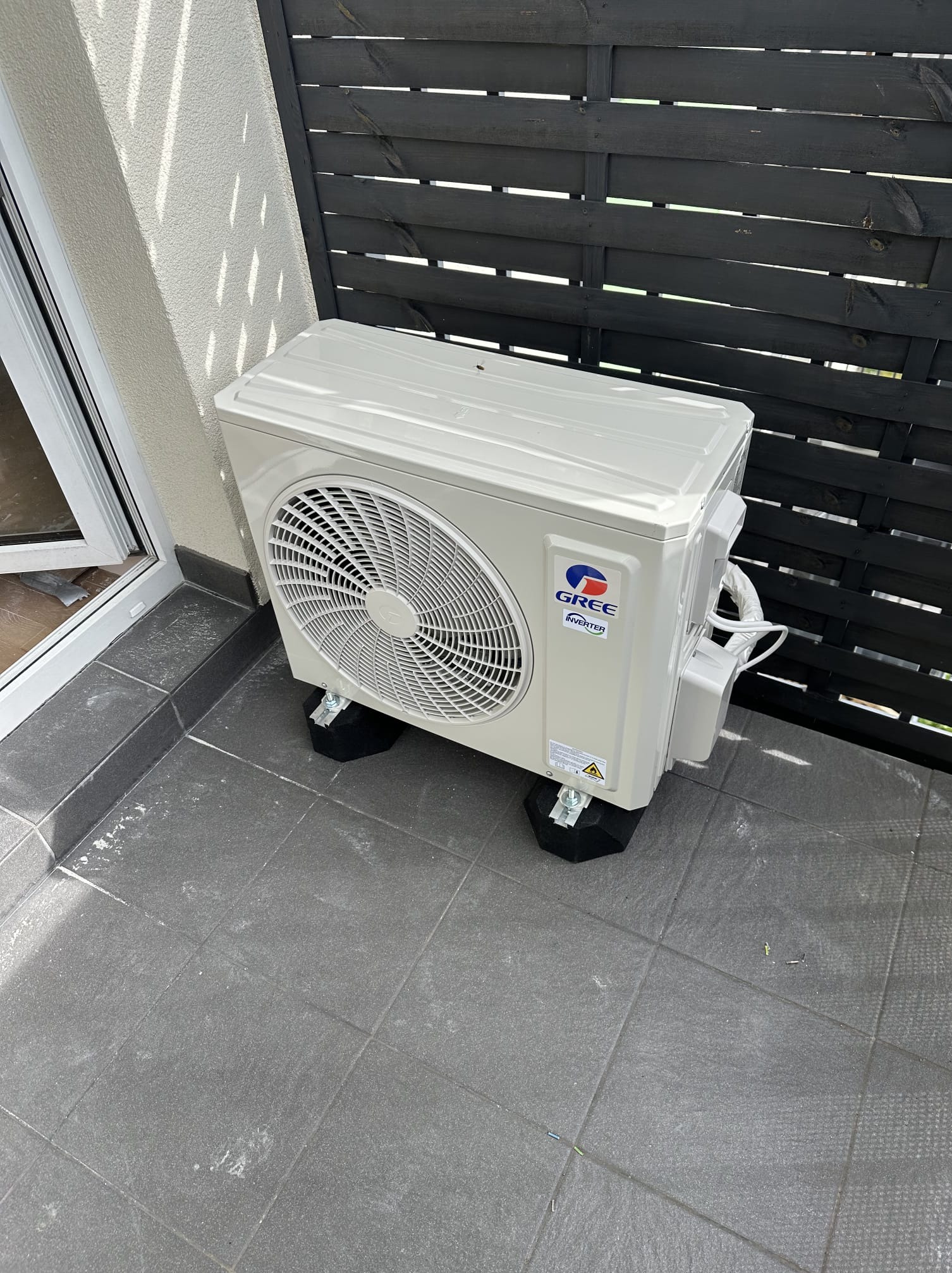 Biała jednostka zewnętrzna klimatyzacji Gree Inverter na szarym balkonie z czarnym drewnianym ogrodzeniem w tle. Widoczne czarne podkładki pod urządzeniem.