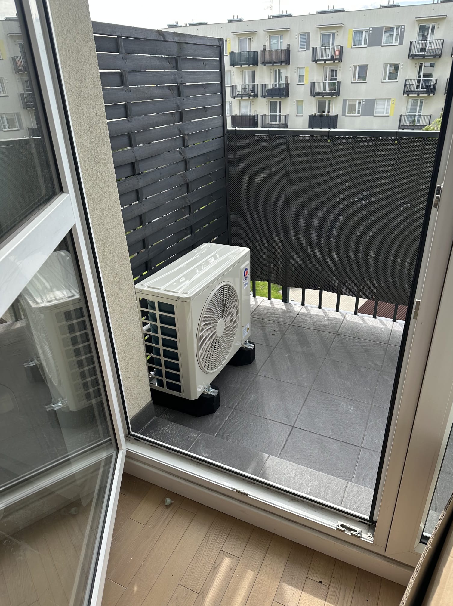 Biała jednostka zewnętrzna klimatyzacji Gree na balkonie z szarą podłogą i czarnym ażurowym ogrodzeniem. Widok z wnętrza przez otwarte drzwi balkonowe.