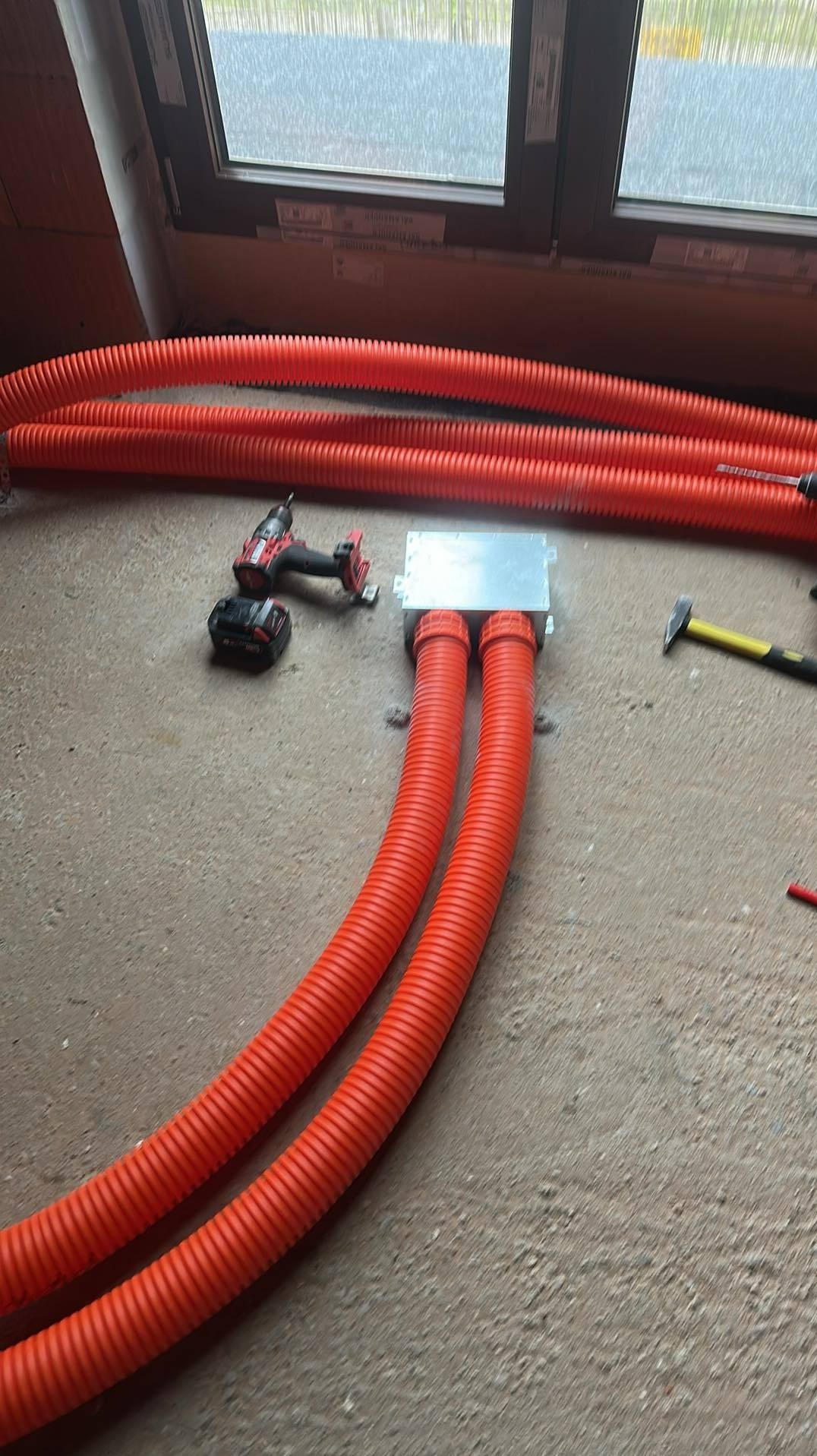 Montaż instalacji ogrzewania podłogowego w Warszawie: pomarańczowe rury w peszlu, narzędzia pracy i okno w tle. Etap prac przed wylaniem posadzki.