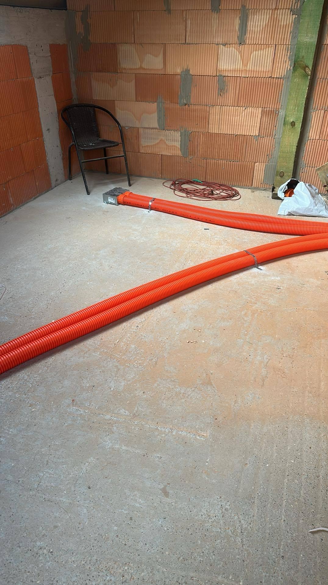 Pomarańczowe rury karbowane ułożone na surowym betonie w pomieszczeniu z ceglanymi ścianami. Widoczny fragment instalacji i krzesło w tle.