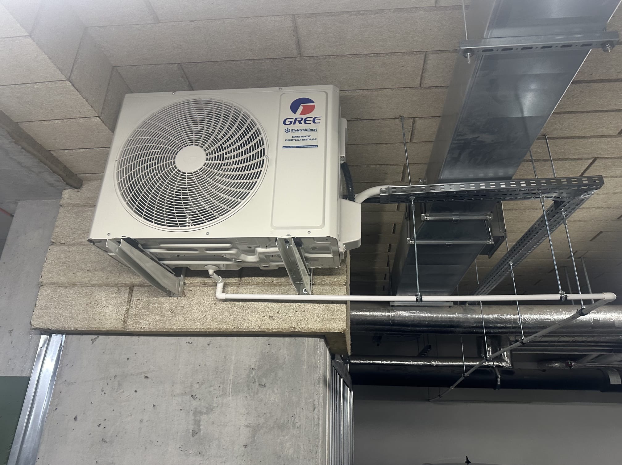 Biała jednostka zewnętrzna klimatyzacji Gree zamontowana na szarej ścianie z betonu komórkowego, widoczne rury i kanały wentylacyjne pod sufitem.