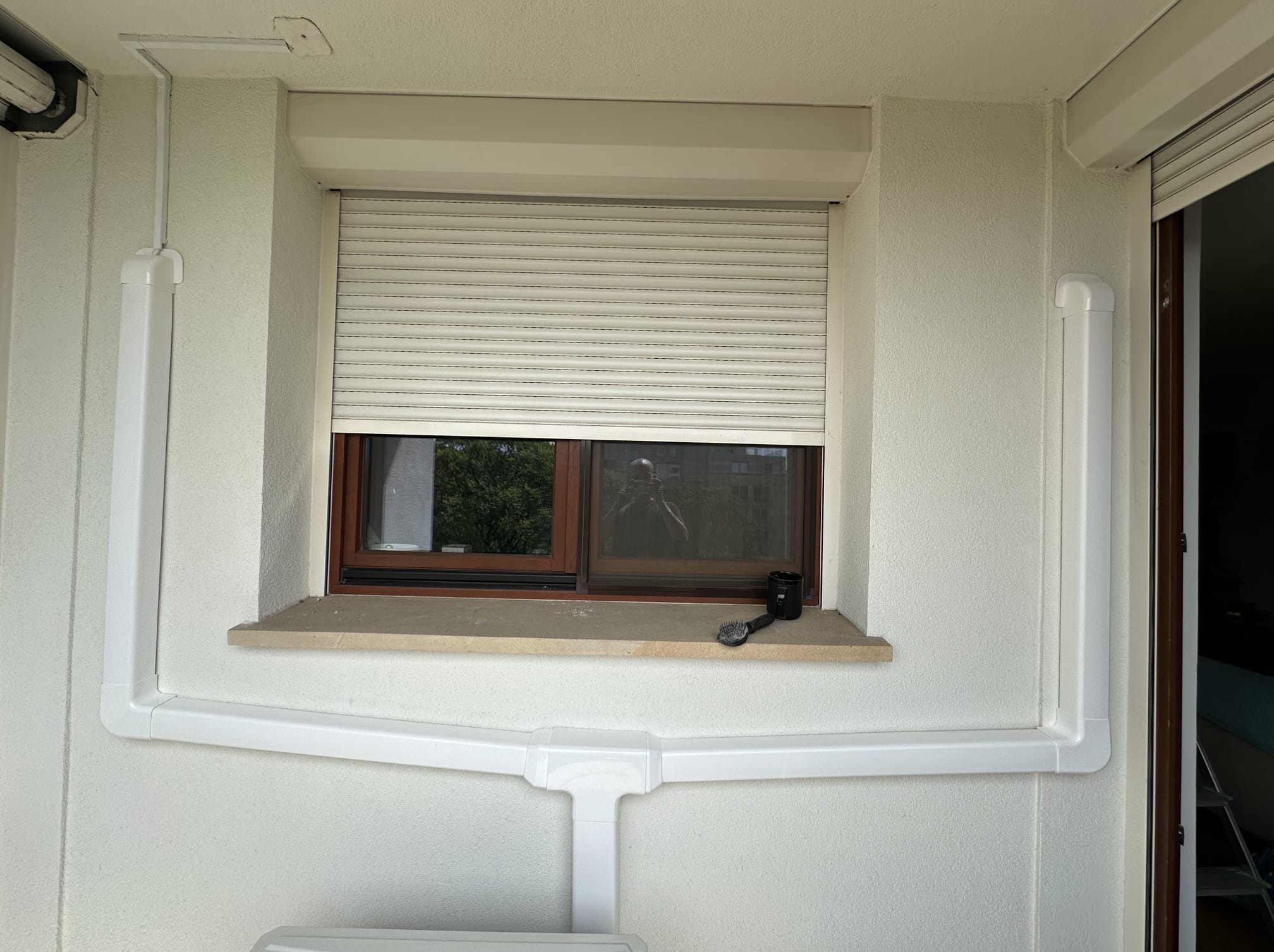 Białe rury instalacyjne na elewacji budynku, prowadzone wzdłuż okna z roletą zewnętrzną. Widoczne elementy mocujące i załamania rur. Na parapecie kubek i szczotka.