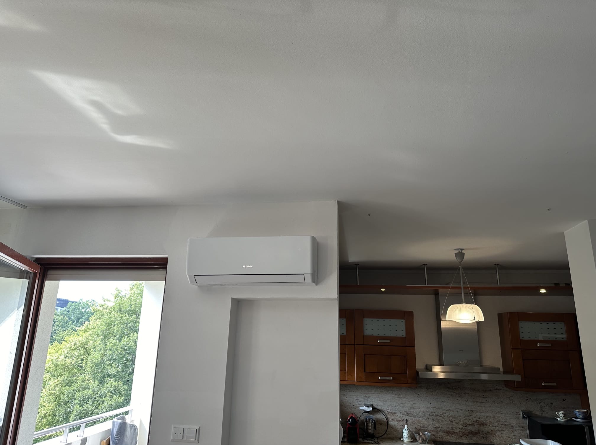 Biały klimatyzator ścienny marki Gree zamontowany w jasnym wnętrzu mieszkania, widok z boku, obok okno balkonowe i fragment zabudowy kuchennej.