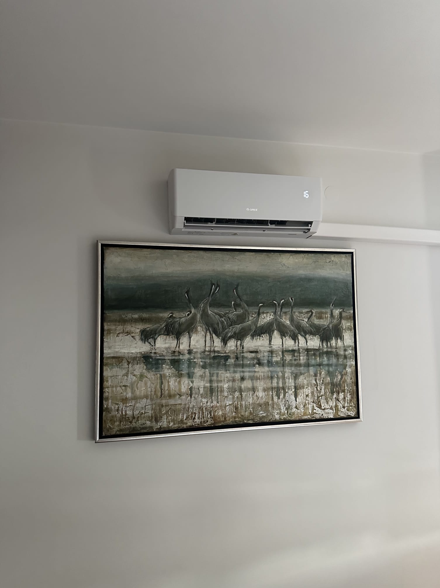 Biała klimatyzacja marki Gree zamontowana nad obrazem z motywem żurawi w srebrnej ramie na jasnej ścianie. Widoczny wyświetlacz temperatury.