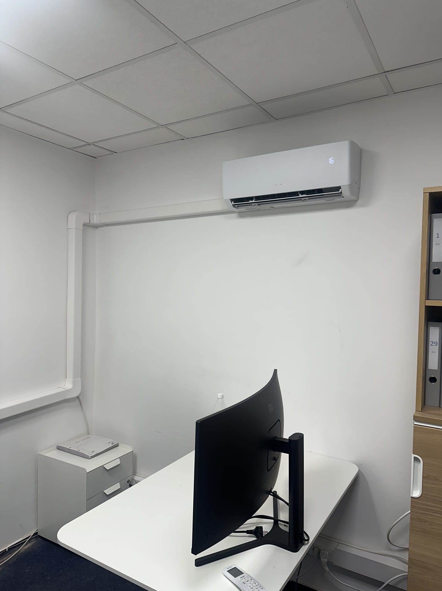 Biały klimatyzator ścienny zamontowany w biurze nad biurkiem z monitorem. Widoczne białe rury instalacyjne i fragment regału z segregatorami.