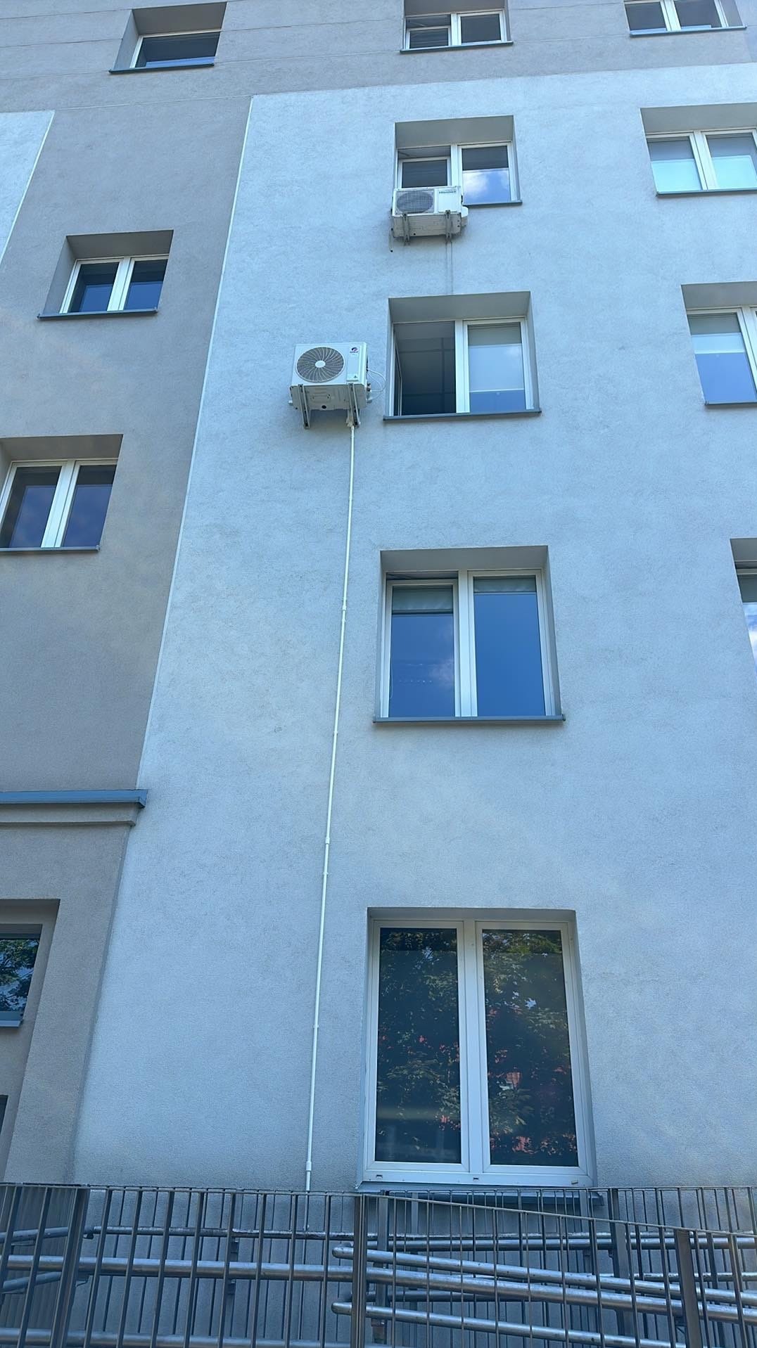 Dwa klimatyzatory zamontowane na elewacji budynku wielorodzinnego, widoczne przewody instalacyjne. Jeden klimatyzator umieszczony nad oknem z otwartym skrzydłem.