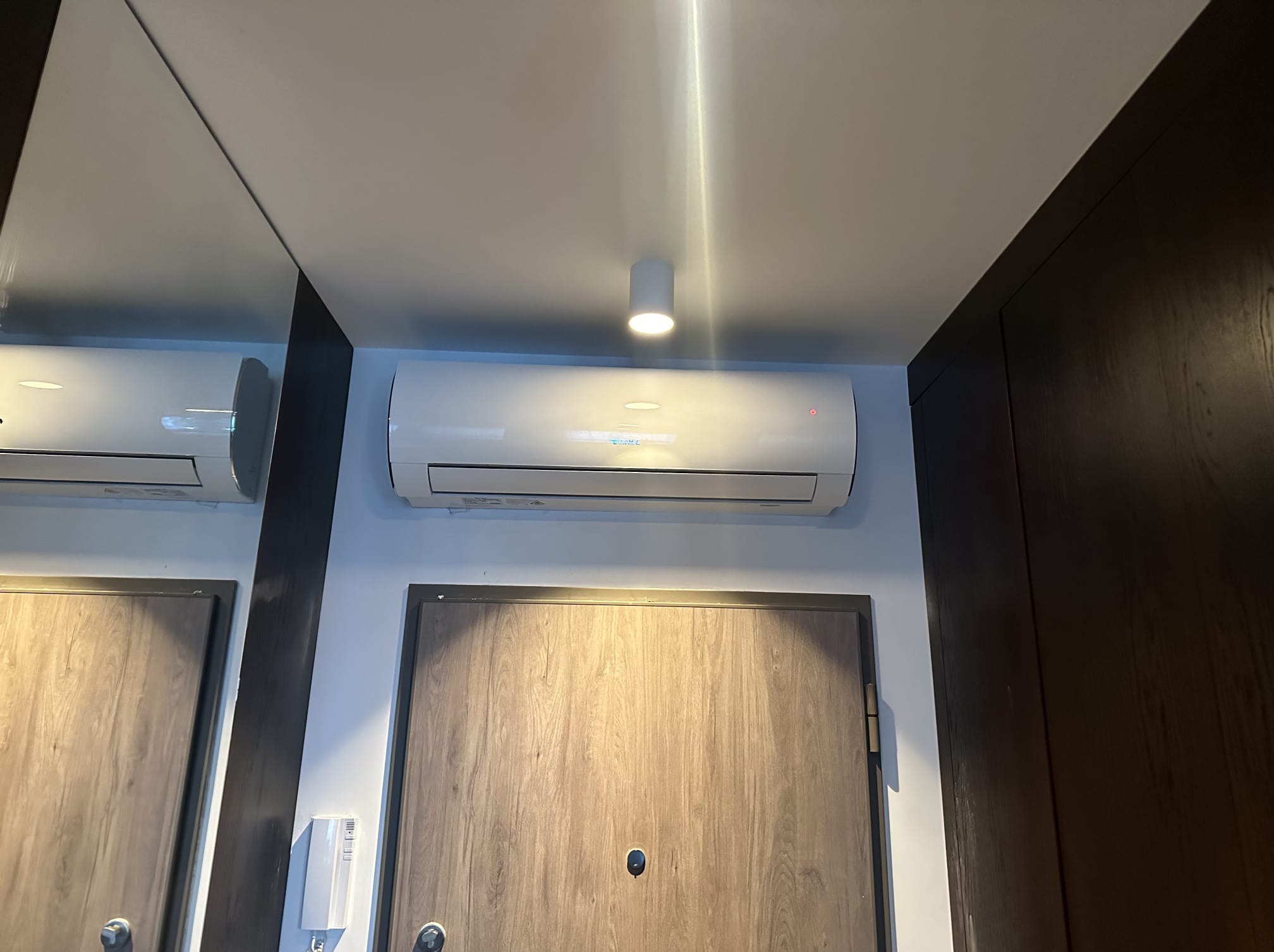 Biały klimatyzator ścienny zamontowany nad drzwiami wejściowymi w mieszkaniu, oświetlony punktowym światłem sufitowym. Widoczne drzwi i fragment lustra.