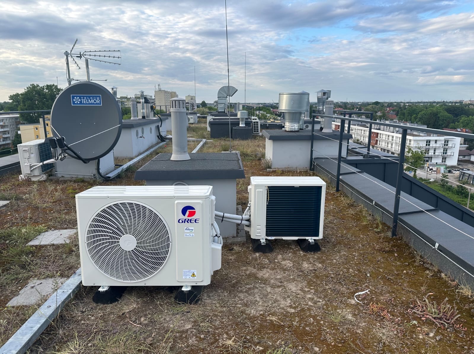 Dwa klimatyzatory GREE na dachu budynku, z anteną satelitarną Telmor w tle i widokiem na miasto. Instalacja klimatyzacji na dachu z zielonym poszyciem.