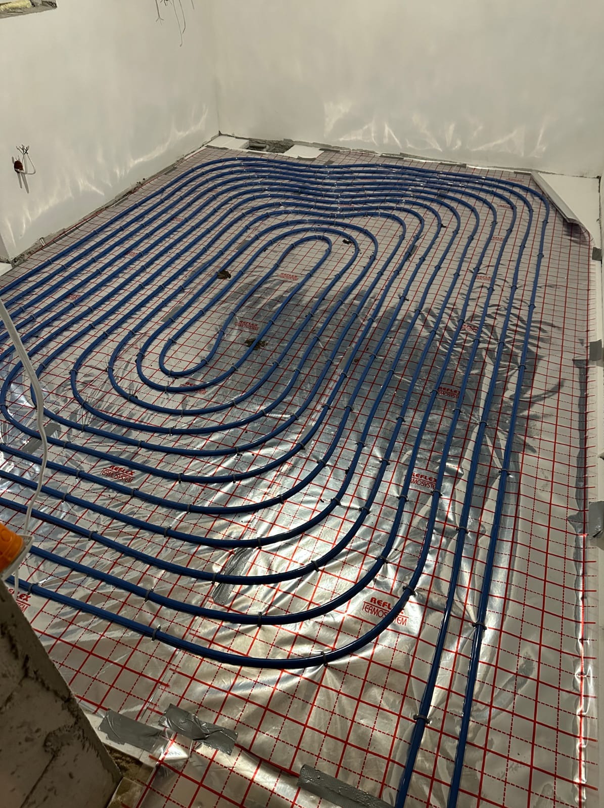 Niebieskie rury ogrzewania podłogowego ułożone spiralnie na srebrnej folii z czerwoną siatką w jasnym pomieszczeniu. Widoczne fragmenty ścian i podłoża w trakcie instalacji.