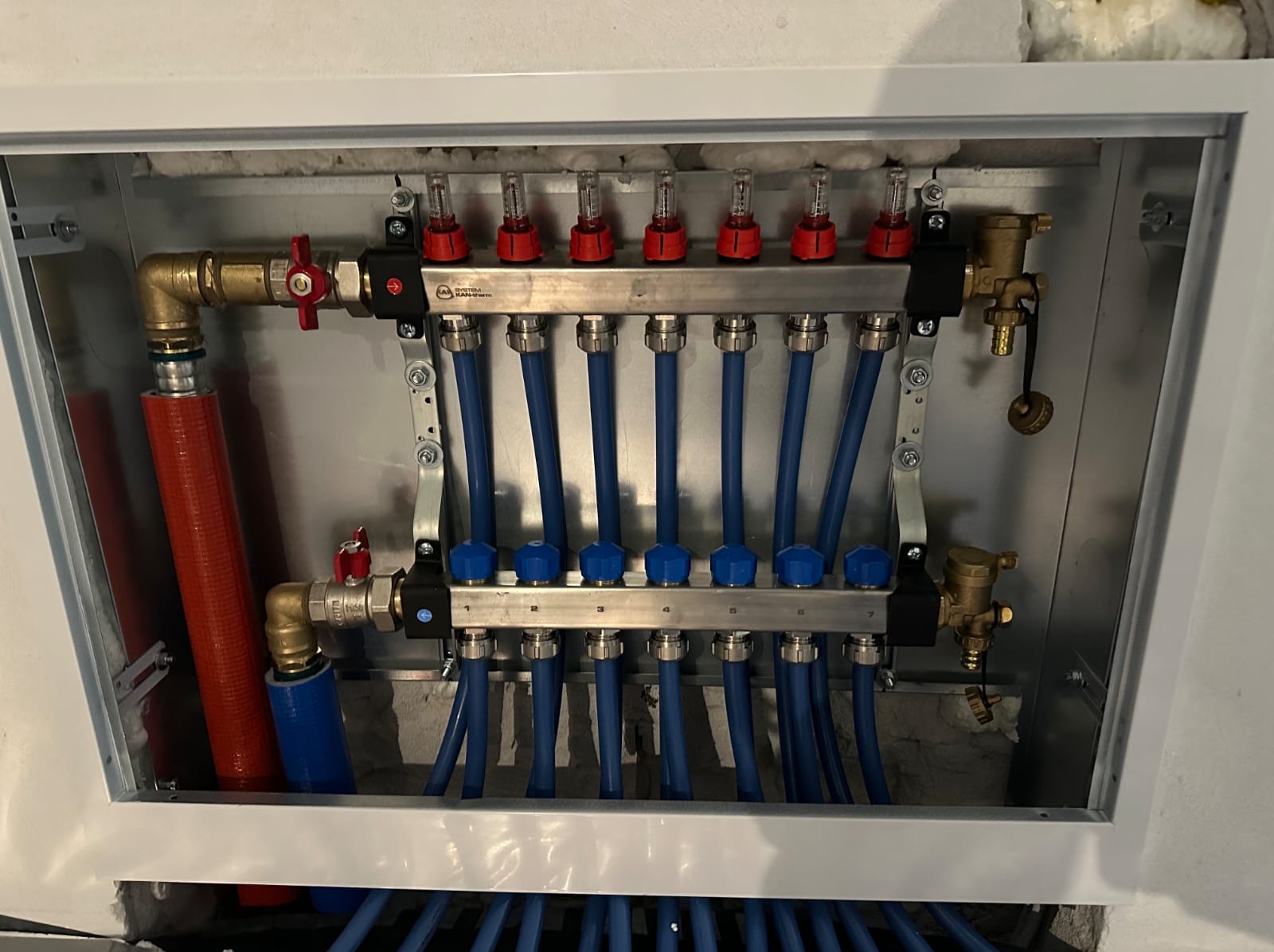 Rozdzielacz ogrzewania podłogowego w metalowej szafce, z widocznymi niebieskimi rurami i elementami regulacyjnymi. Widok z góry, instalacja w trakcie montażu.