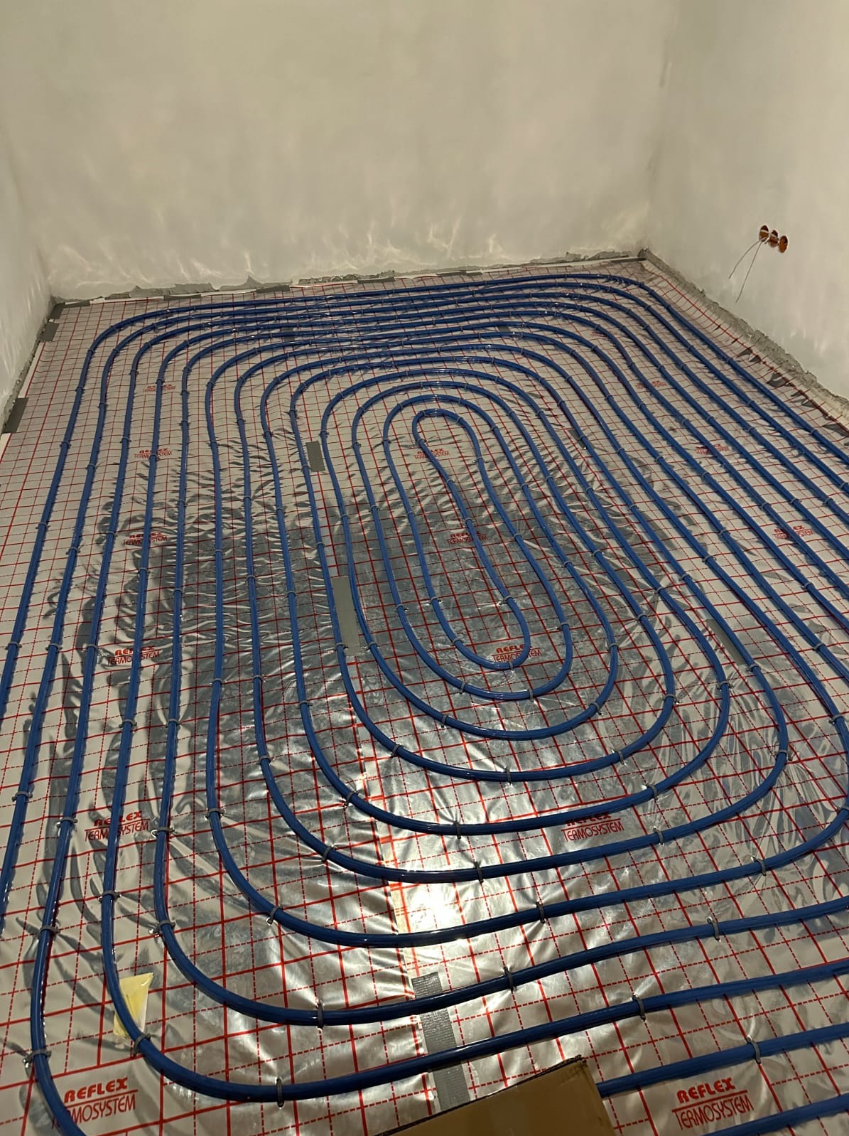 Niebieskie rury ogrzewania podłogowego ułożone spiralnie na srebrnej folii z czerwoną kratką w surowym wnętrzu. Widoczne mocowania rur i fragment kartonu w rogu.