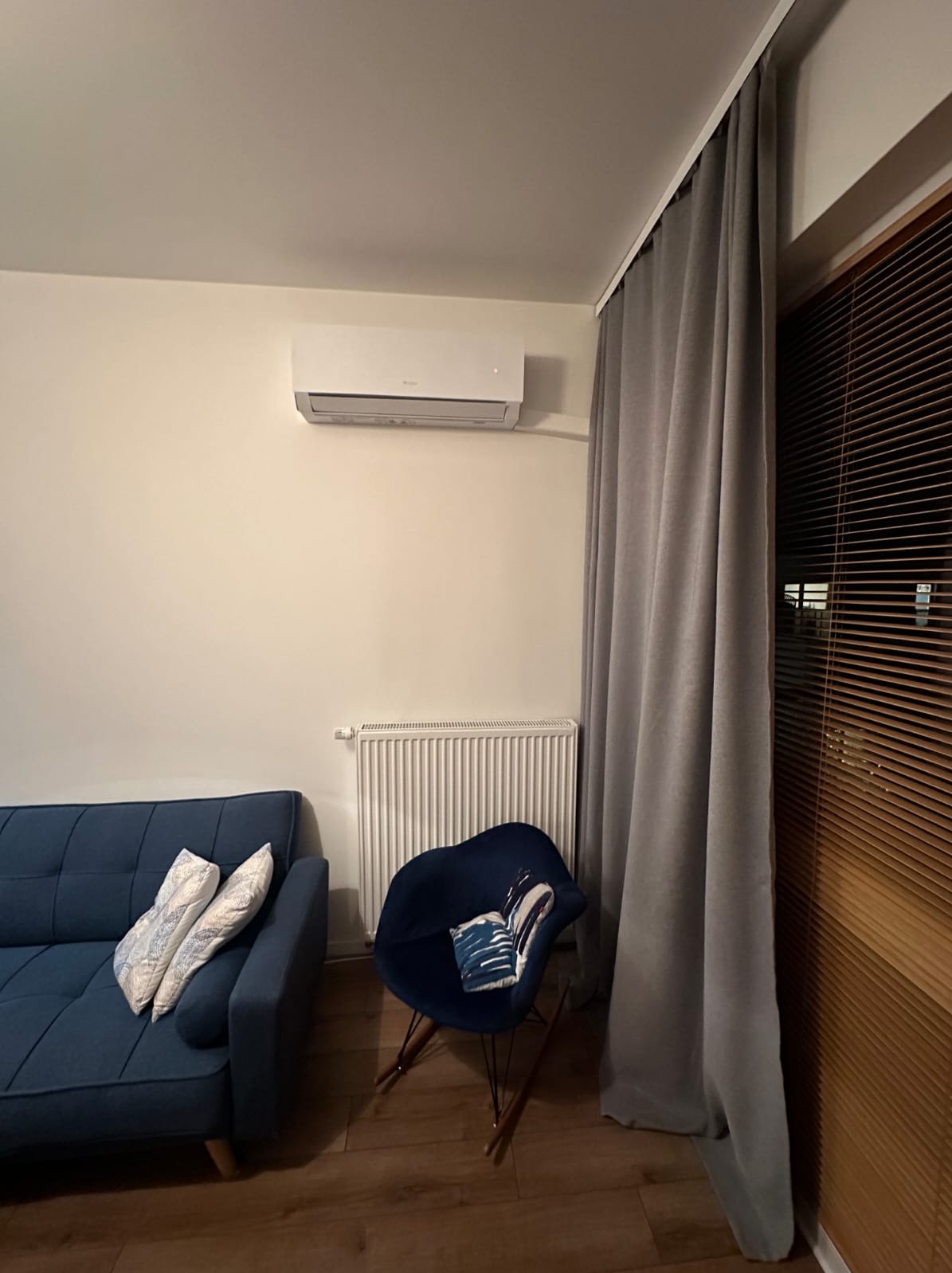 Biały klimatyzator ścienny w salonie z niebieską sofą i grzejnikiem. Widoczne zasłony i żaluzje w oknie. Ustawienie mebli w pokoju dziennym.