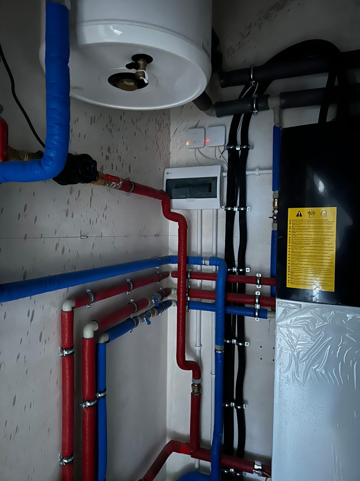 Złożona instalacja hydrauliczna z rurami w izolacji (czerwone i niebieskie) oraz czarnymi rurami, zbiornik i biała skrzynka elektryczna na tle ściany.