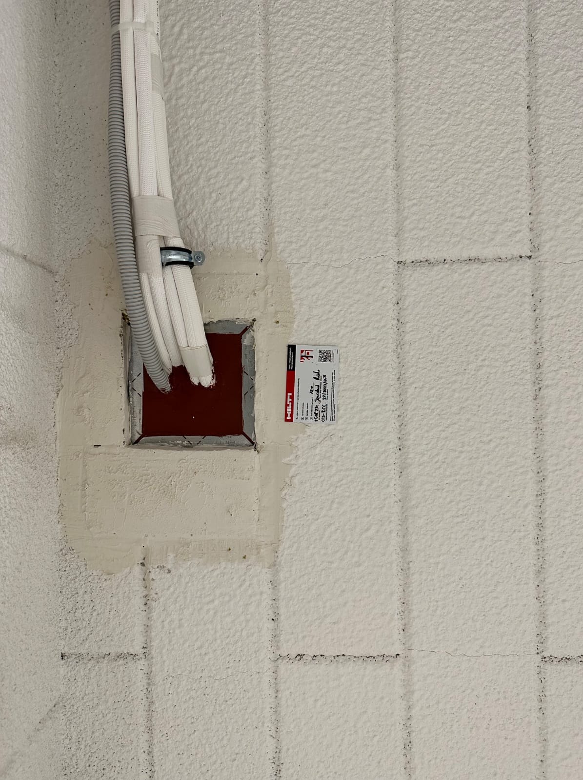 Przewody wentylacyjne w izolowanej ścianie, zabezpieczone w otworze. Widoczna etykieta Hilti. Biała, teksturowana powierzchnia ściany z widocznymi liniami podziału.
