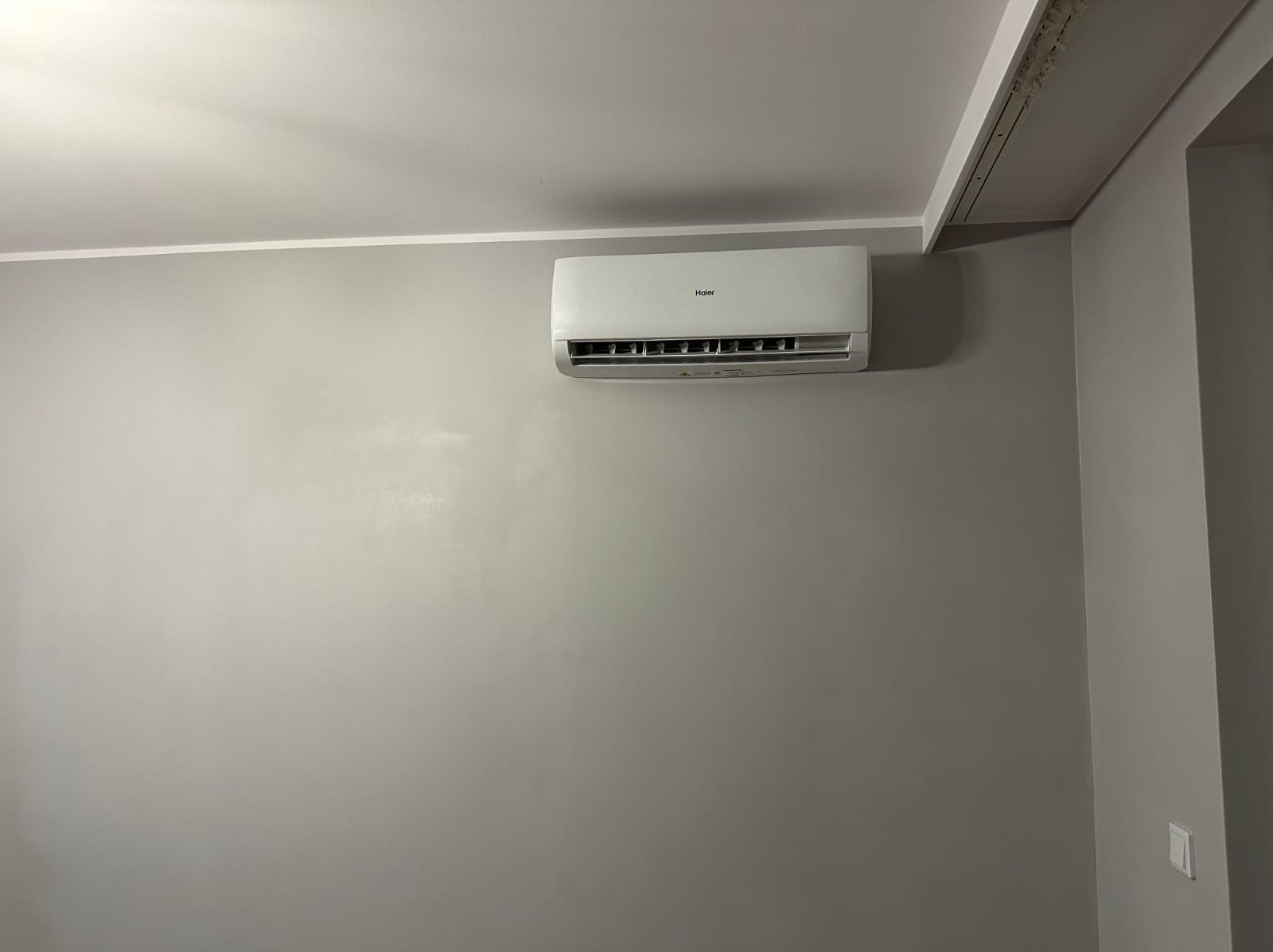 Biały klimatyzator Haier zamontowany na szarej ścianie w pomieszczeniu. Widoczny fragment sufitu z listwą. Minimalistyczny design wnętrza.