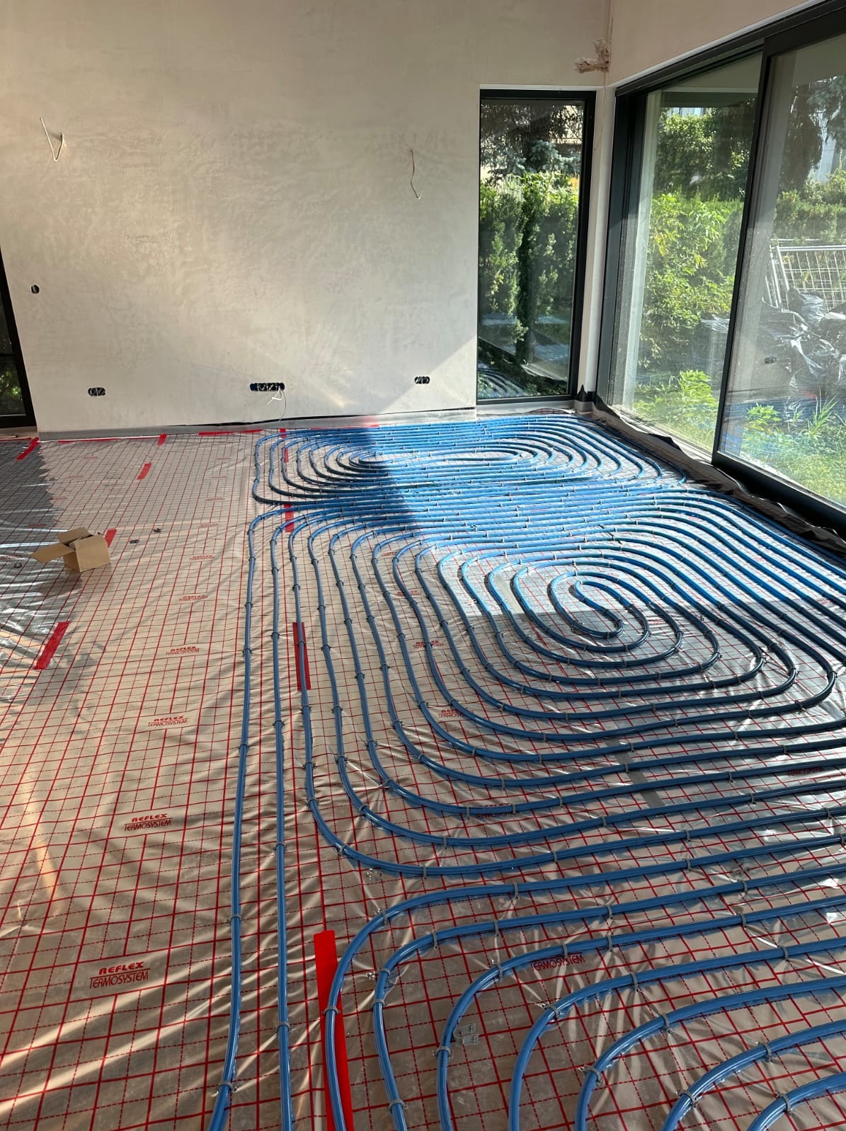 Niebieskie rury ogrzewania podłogowego ułożone na folii z czerwoną siatką w jasnym pomieszczeniu z dużymi oknami. Widoczne elementy instalacji elektrycznej na ścianie.