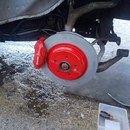 Kaspro Michał Kasprowski - Czerwony zacisk Brembo i tarcza hamulcowa po renowacji na tle zawieszenia pojazdu. Widoczne narzędzia i podnośnik. Detal z warsztatu samochodowego.