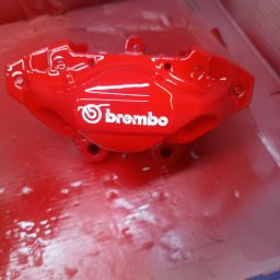 Kaspro Michał Kasprowski - Czerwony zacisk hamulcowy Brembo po renowacji, świeżo pomalowany, z widocznym logo, leżący w kartonowym pudełku. Detal z warsztatu samochodowego.