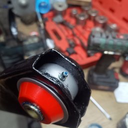 Kaspro Michał Kasprowski - Wahacz samochodowy z nową, czerwoną tuleją poliuretanową i smarowaniem, gotowy do montażu. W tle narzędzia warsztatowe i skrzynka z nasadkami.