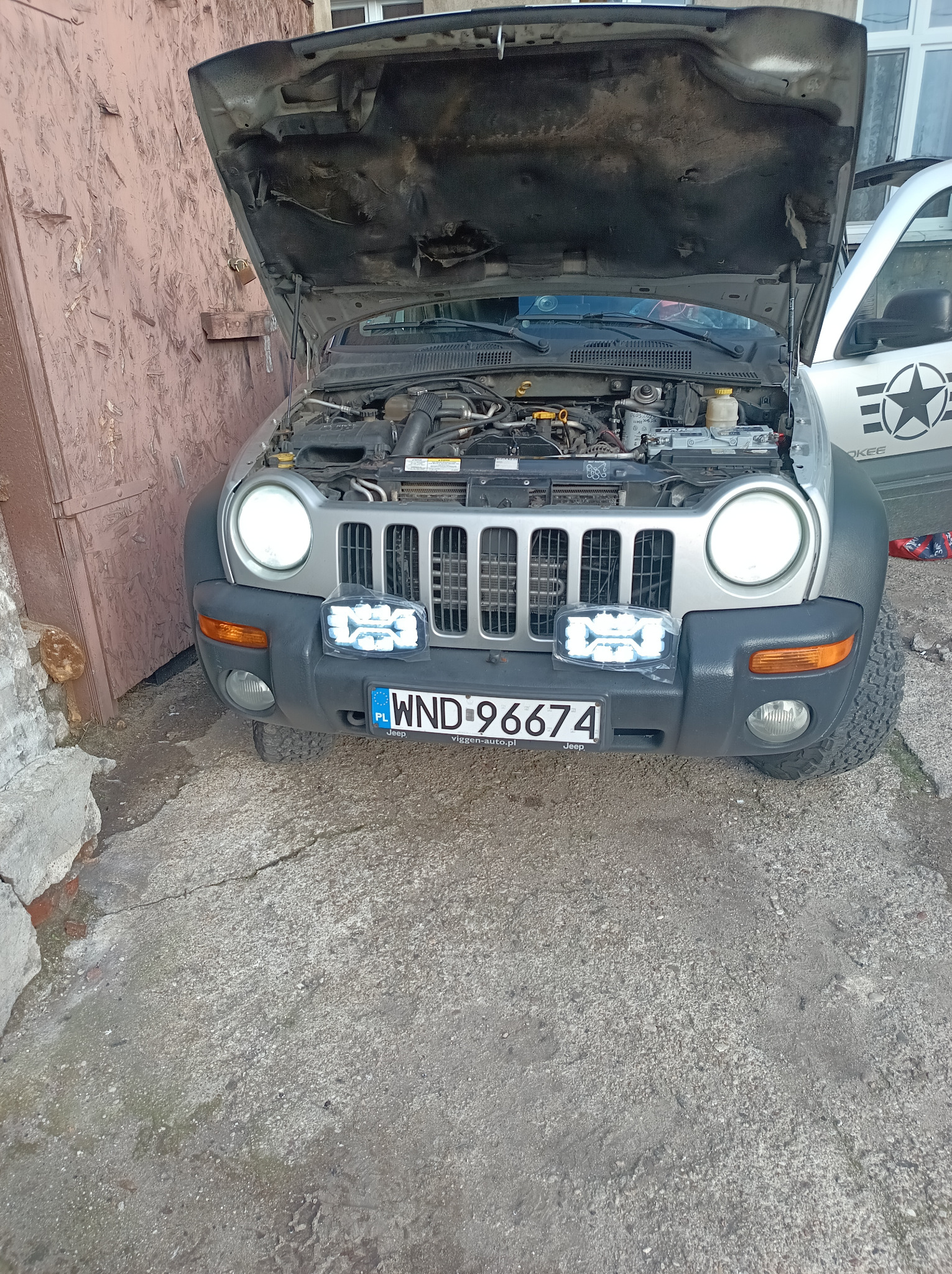 Srebrny Jeep z otwartą maską, widoczne elementy silnika i nowe, jasne reflektory LED zamontowane z przodu. Tablica rejestracyjna z przodu pojazdu.