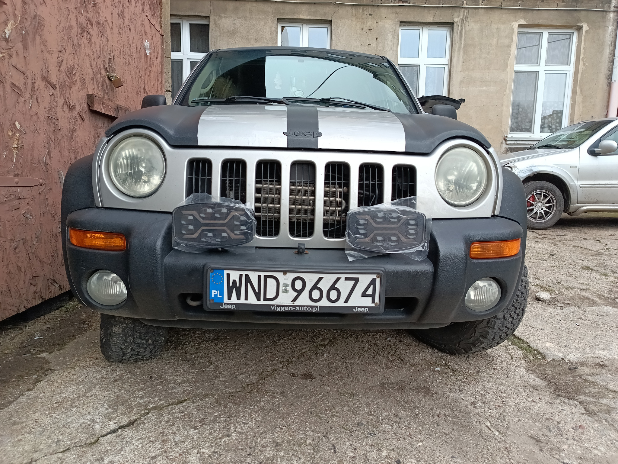 Srebrny Jeep Liberty z czarnym pasem na masce, tablicą rejestracyjną WND 96674 i dodatkowymi światłami LED przed grillem, zaparkowany na zewnątrz budynku.