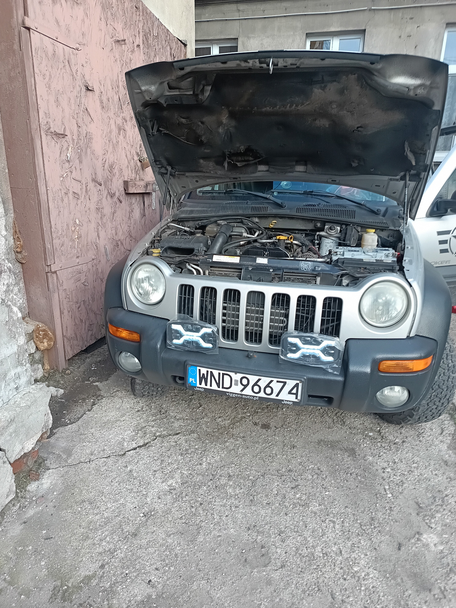 Srebrny Jeep z otwartą maską, widoczny silnik. Na zderzaku leży nowa lampa LED w opakowaniu. Tablica rejestracyjna WND 96674. Tło: brama i budynek.