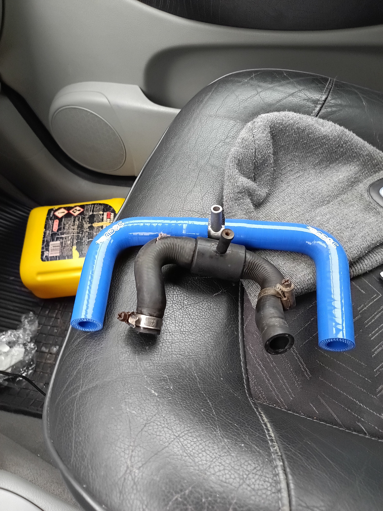Dwa węże samochodowe: nowy, niebieski i stary, czarny, leżą na czarnym skórzanym siedzeniu auta obok żółtego pojemnika z płynem. Węże z metalowymi zaciskami.