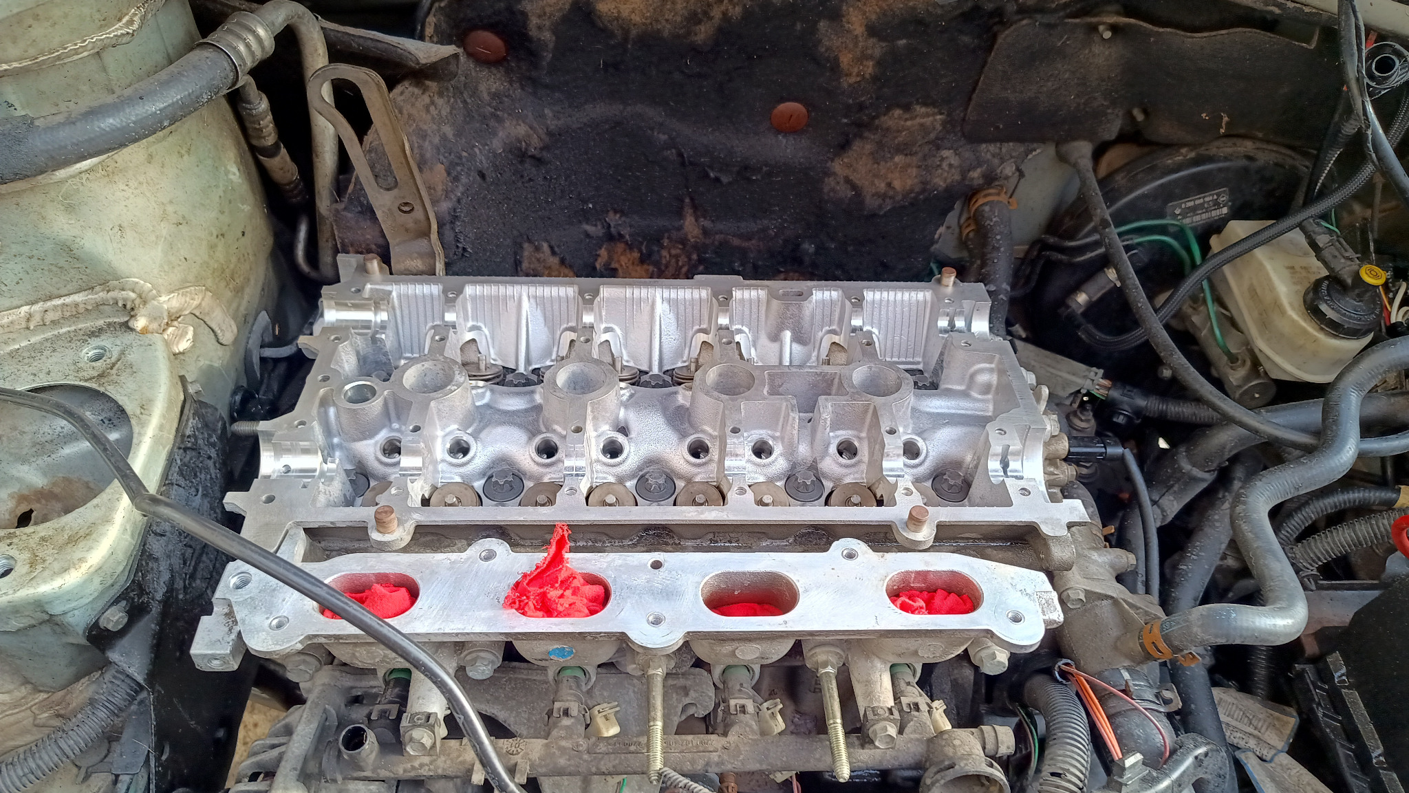 Silnik samochodowy w trakcie naprawy: widoczna głowica cylindrów z czerwonymi zabezpieczeniami w otworach, przewody i elementy osprzętu. Detal z warsztatu.