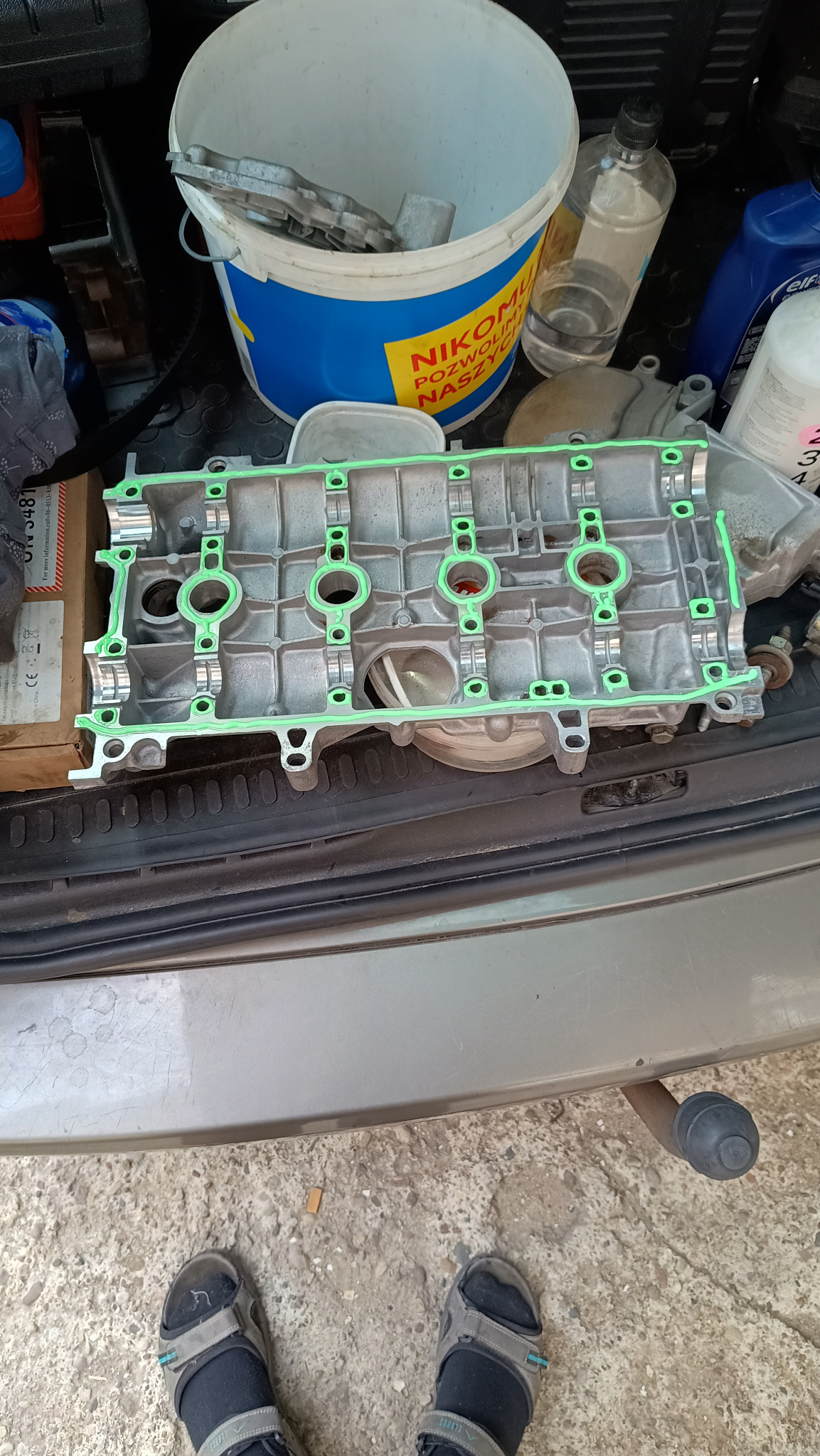Głowica silnika z nową, zieloną uszczelką, leżąca na bagażniku samochodu. Widoczne narzędzia i płyny eksploatacyjne w tle.