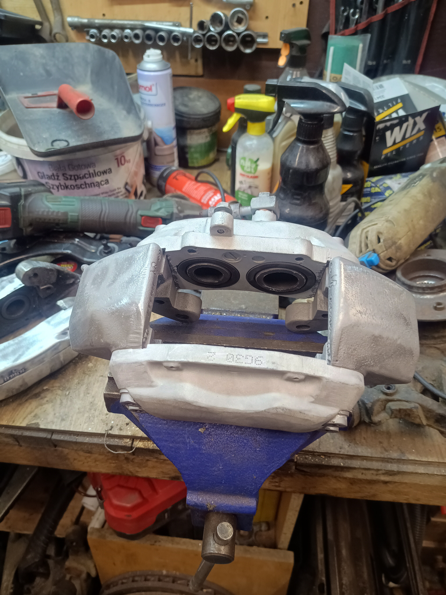 Szary zacisk hamulcowy w imadle na warsztacie, po renowacji. Widoczne narzędzia i akcesoria warsztatowe w tle. Detal po naprawie w warsztacie samochodowym.