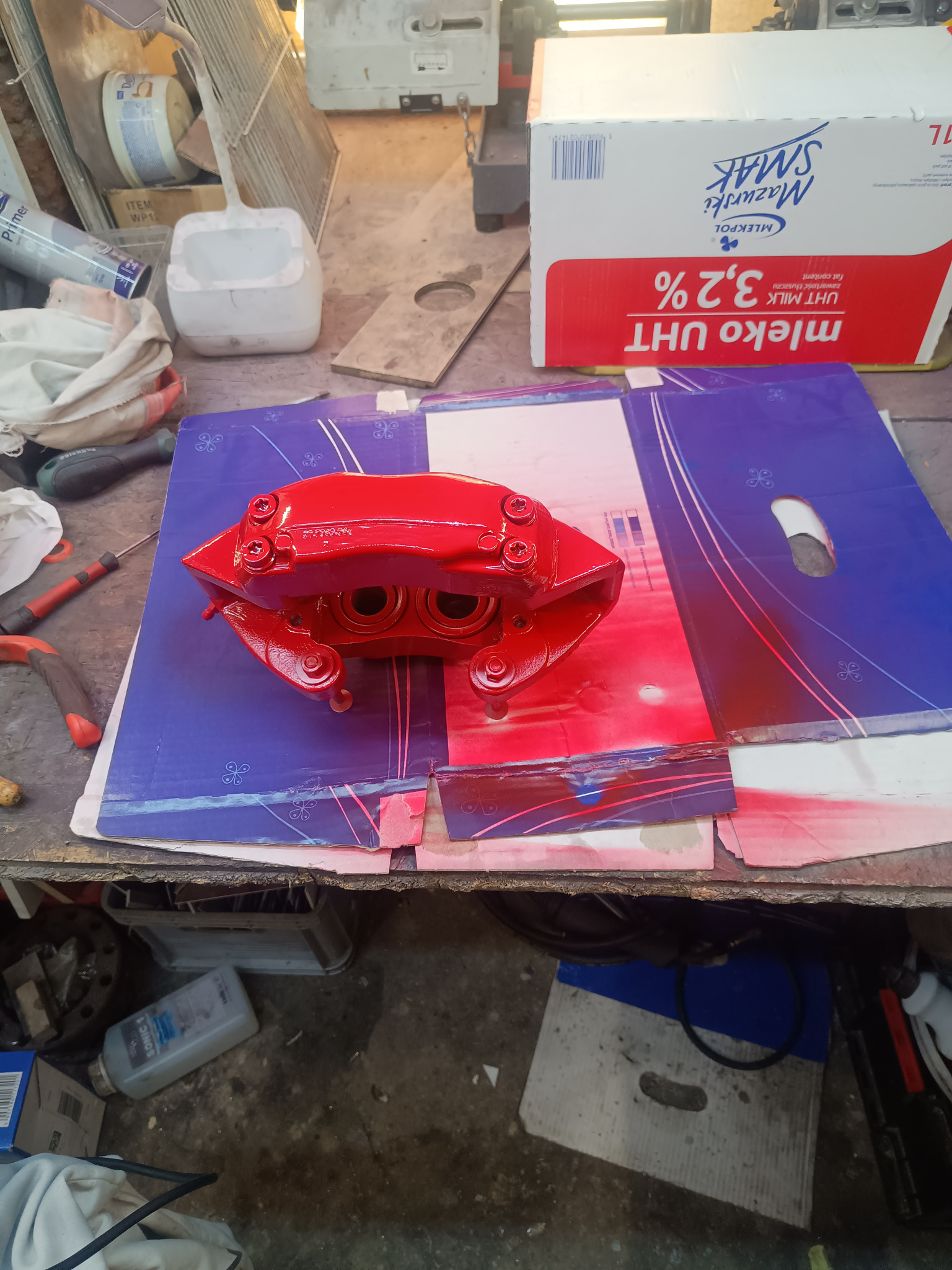 Czerwony, pomalowany zacisk hamulcowy na kartonie z niebieskim wzorem. Widoczne ślady farby i narzędzia. W tle karton mleka i inne przedmioty warsztatowe.