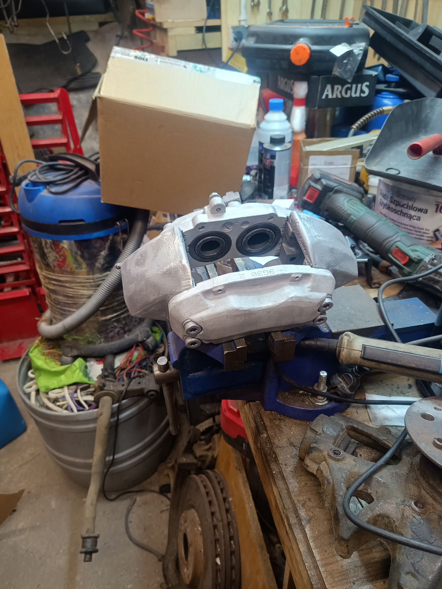 Szary zacisk hamulcowy w imadle, po renowacji, na warsztatowym stole. W tle narzędzia i akcesoria samochodowe. Widoczne ślady pracy.