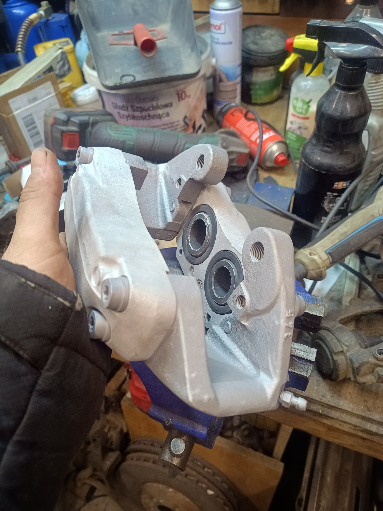 Szary, odnowiony zacisk hamulcowy w imadle, trzymany w dłoni. Widoczne ślady pracy warsztatowej i narzędzia w tle. Detal po renowacji.