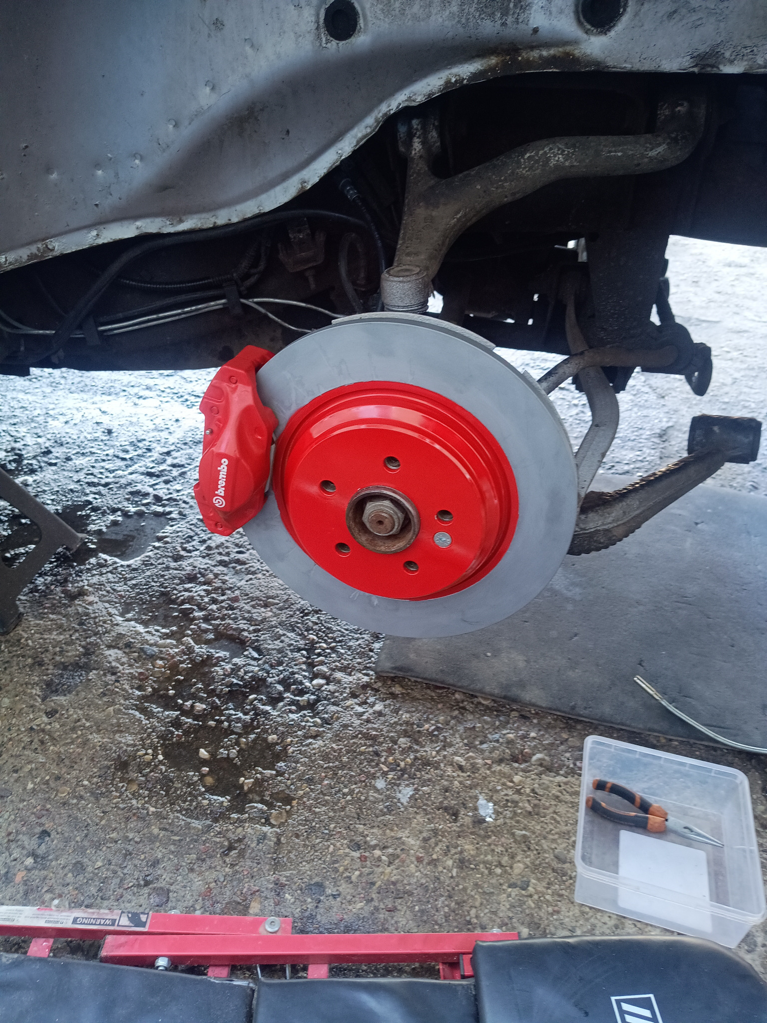 Czerwony zacisk Brembo i tarcza hamulcowa po renowacji na tle zawieszenia pojazdu. Widoczne narzędzia i podnośnik. Detal z warsztatu samochodowego.