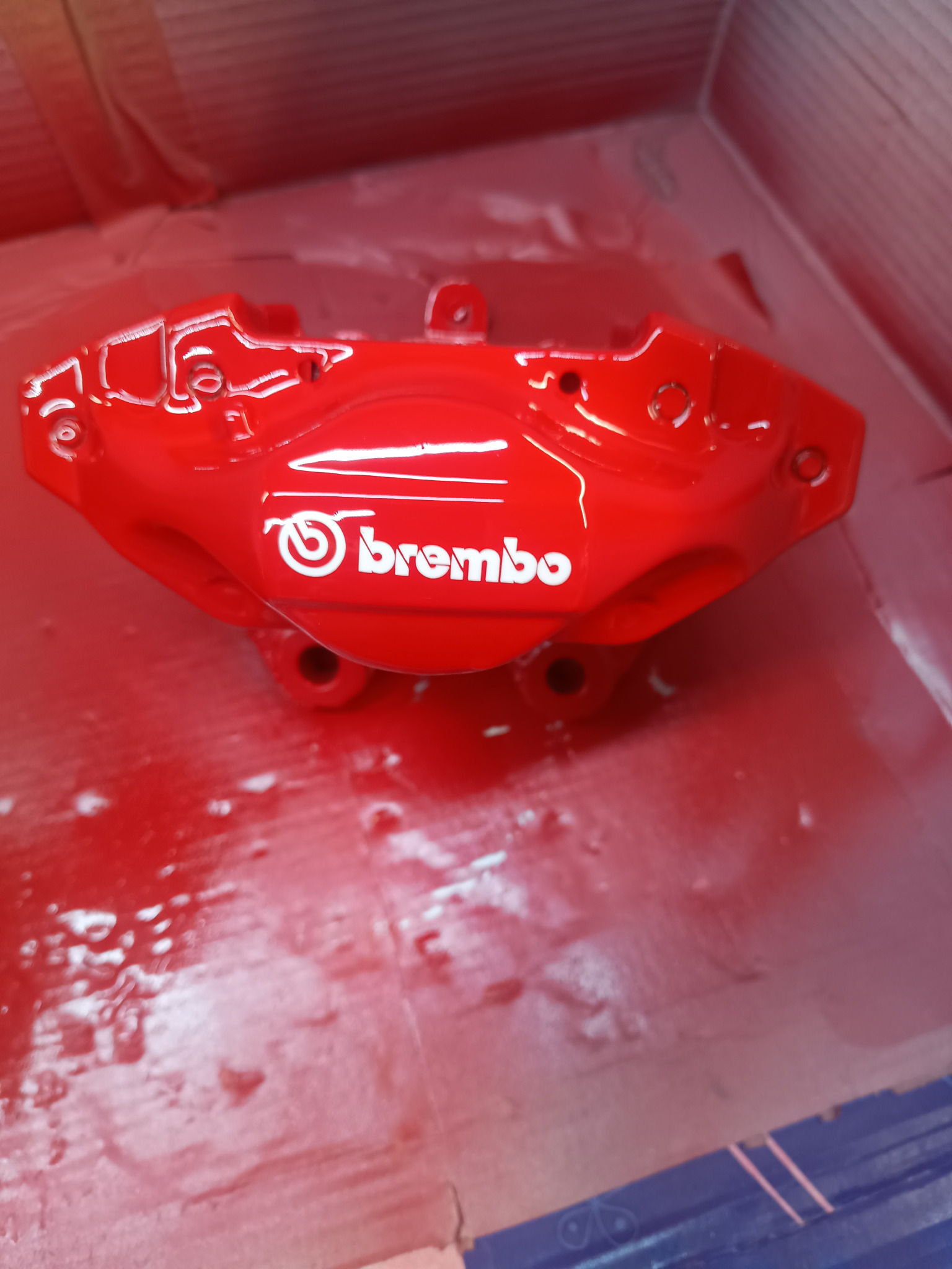 Czerwony zacisk hamulcowy Brembo po renowacji, świeżo pomalowany, z widocznym logo, leżący w kartonowym pudełku. Detal z warsztatu samochodowego.